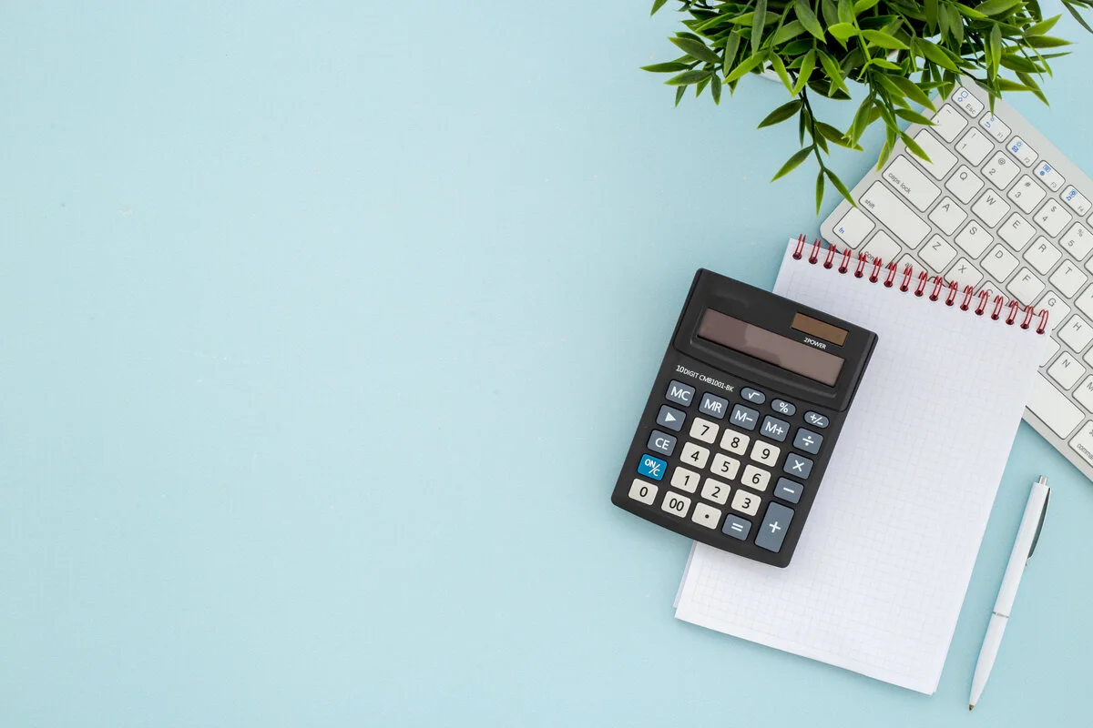 Calculator and notebook on a light blue desk with a keyboard, pen, and plant, representing business tax planning and financial organization.