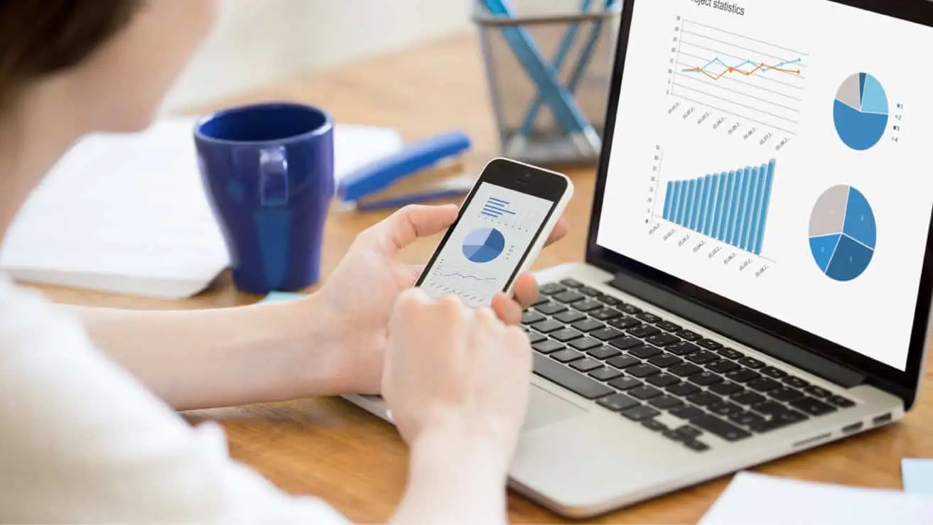 Woman analyzing data using smartphone and laptop with charts displayed on both screens, depicting effective digital analytics strategy.